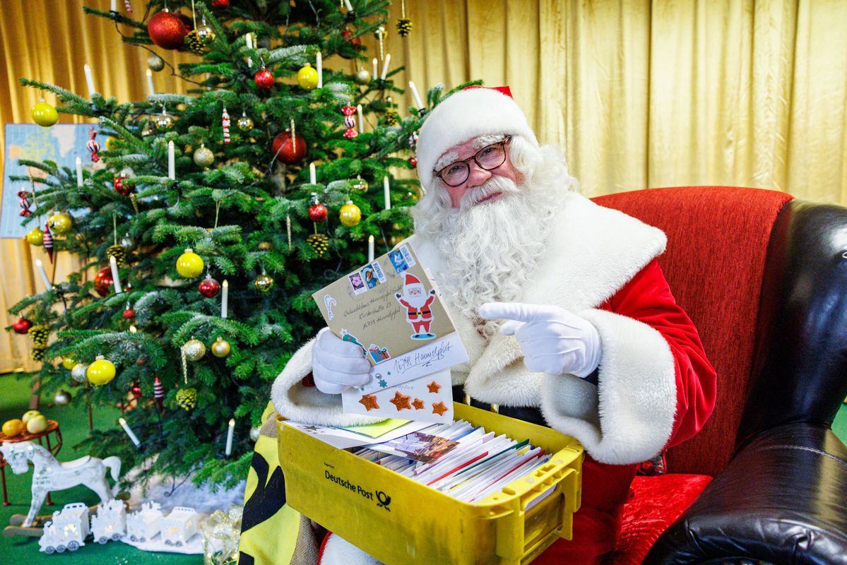 Das Foto zeigt einen Weihnachtsmann in seinem großen roten Sessel vor einem gelben Vorhang sitzend, auf dem Schoss hält er einen großen Weidenkorb, gefüllt mit Weihnachtspostkarten. Er blickt in die Kamera, links hinter ihm steht ein bunt geschmückter Weihnachtsbaum.